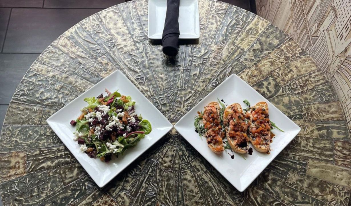 Two plated salads on a round patterned table, with a person’s lower legs visible at the top.