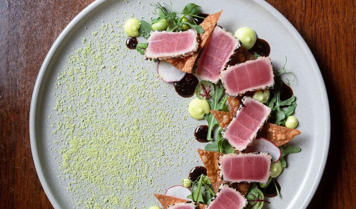 Plated tuna sashimi with greens and vegetables on a white dish, drizzled with sauce