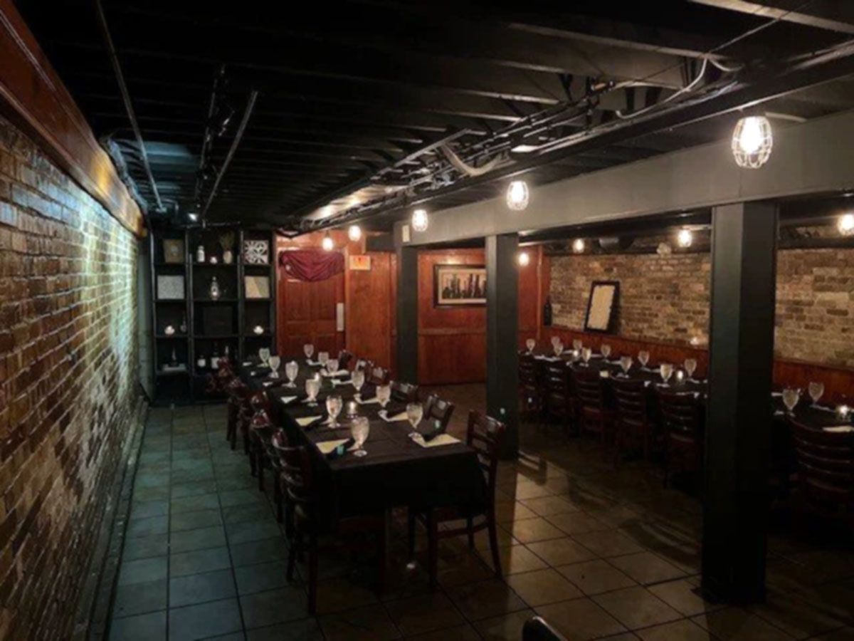 Dimly lit restaurant interior with exposed brick, dark tables, and set dining tables along a narrow aisle