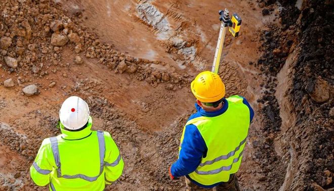 Two construction workers in hard hats and neon vests inspect a muddy excavation site.