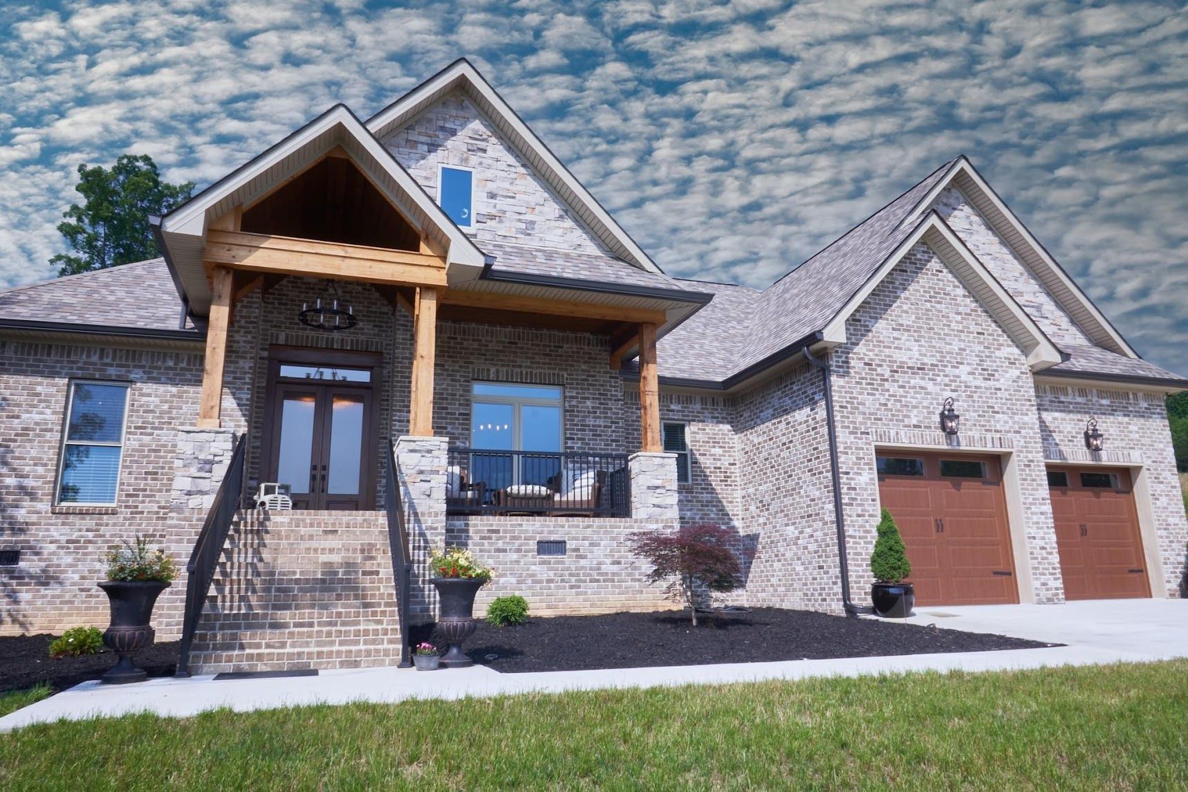 Large stone house with a covered front porch, balcony, and two-car garage under a bright sky