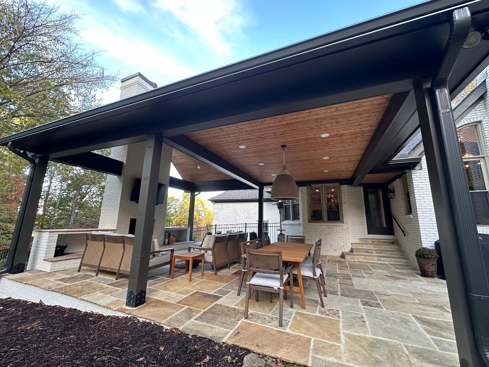 Covered stone patio with outdoor dining set, fireplace, and dark wood ceiling at a house