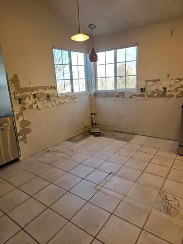 Empty tiled kitchen with peeling walls, two windows, and a hanging light fixture.