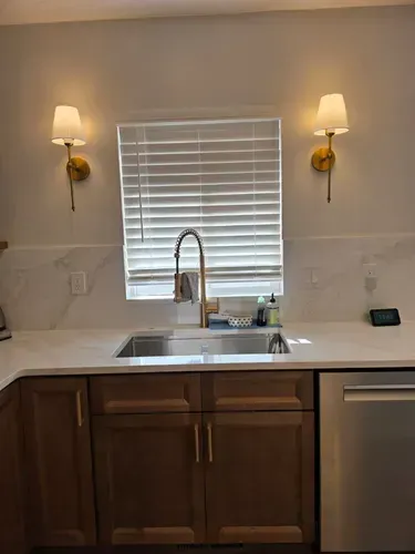 Kitchen sink with white countertops, wooden cabinets, and wall sconces beside a window with blinds.