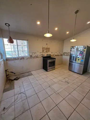 Empty tiled kitchen with stainless refrigerator, stove, pendant lights, and a window.