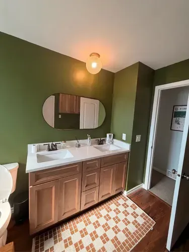 Small bathroom with green walls, double sink vanity, round mirror, and patterned tile floor.