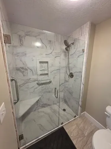 Modern glass walk-in shower with marble tile, built-in shelf, and grab bars in a small bathroom
