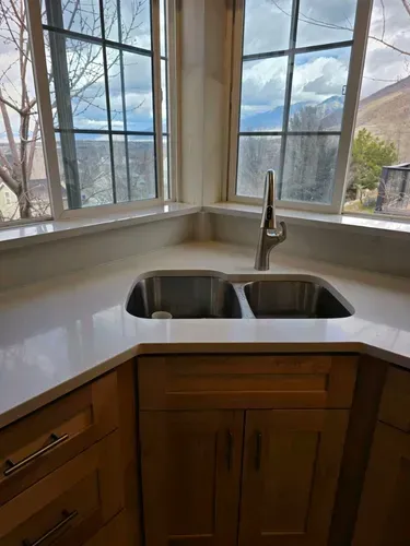 Corner kitchen sink under large windows with a chrome faucet and wooden cabinets.