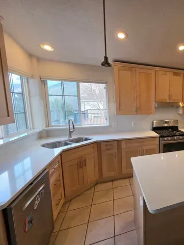 Bright kitchen with wooden cabinets, white countertops, tiled floor, and large windows above the sink.