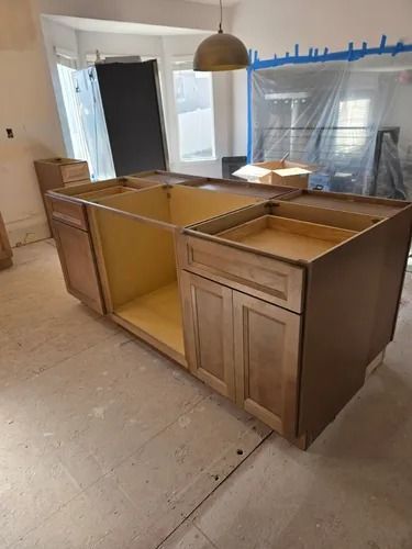 Partially installed wooden kitchen cabinets in an unfinished room with plastic-covered windows.