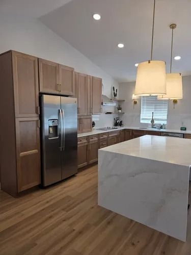 Modern kitchen with wood cabinets, stainless fridge, white island, pendant lights, and wood floor.