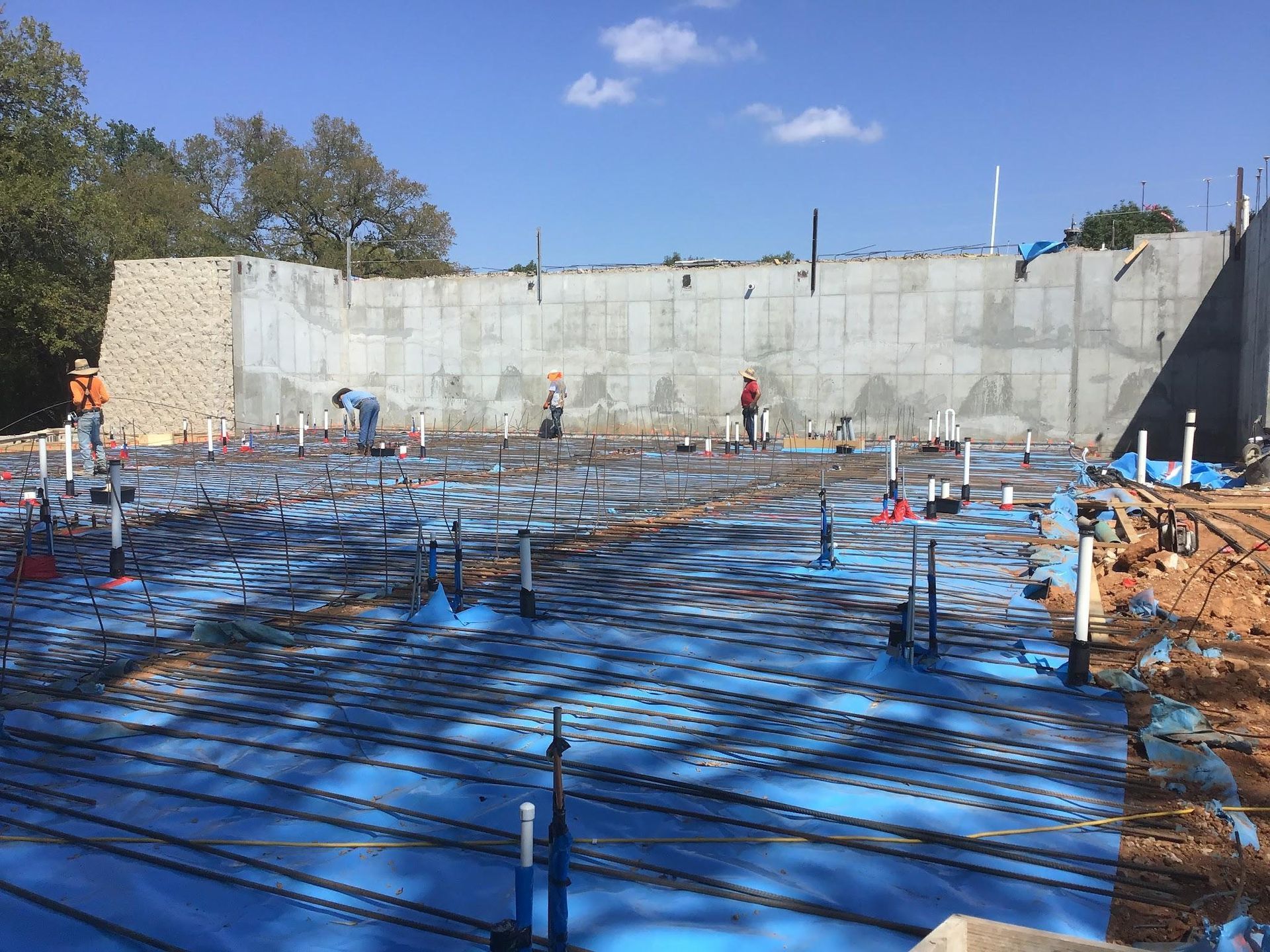 Construction site with workers, concrete walls, rebar, and plumbing, blue underlayment, sunny day.
