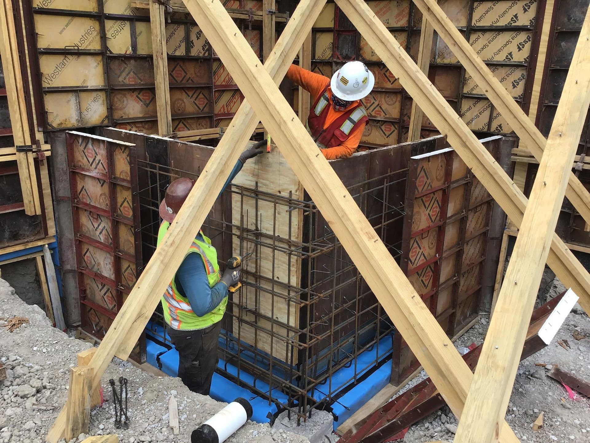 Construction workers pouring concrete into a form. Workers wear safety vests and helmets. Forms are braced with wood.