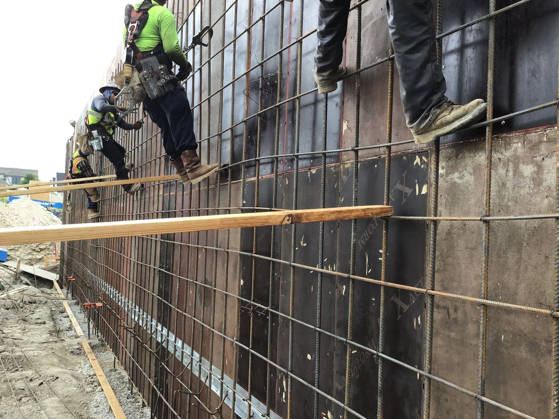 Construction workers on a concrete wall form, wearing safety gear. Black wire mesh, brown plywood, and wood beams are visible.
