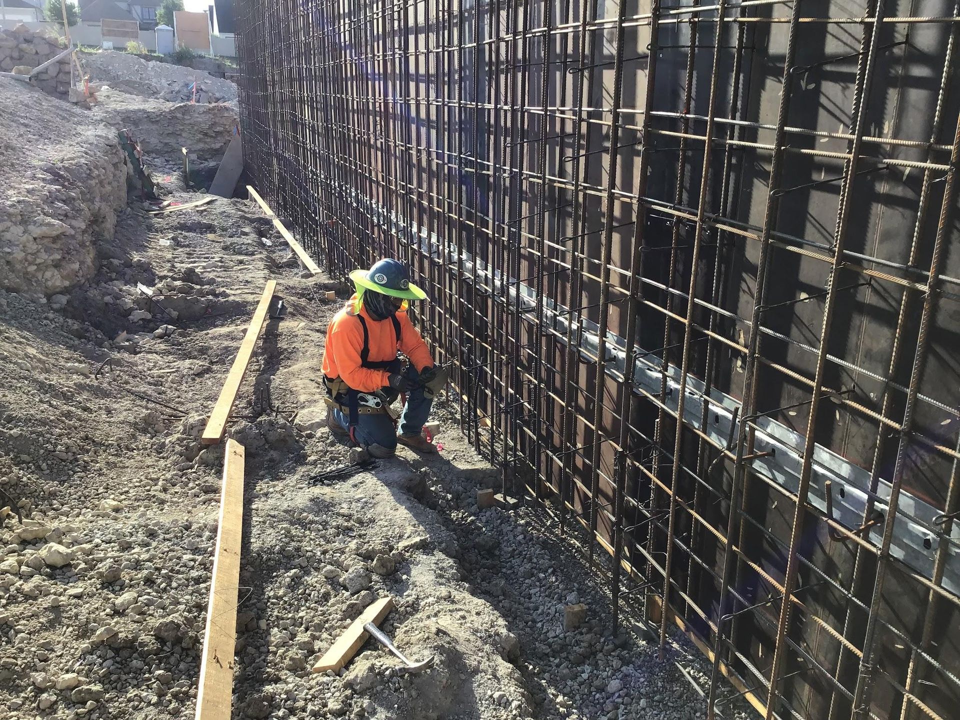 Construction worker kneels near rebar cage, wearing orange vest and hard hat. Construction site.