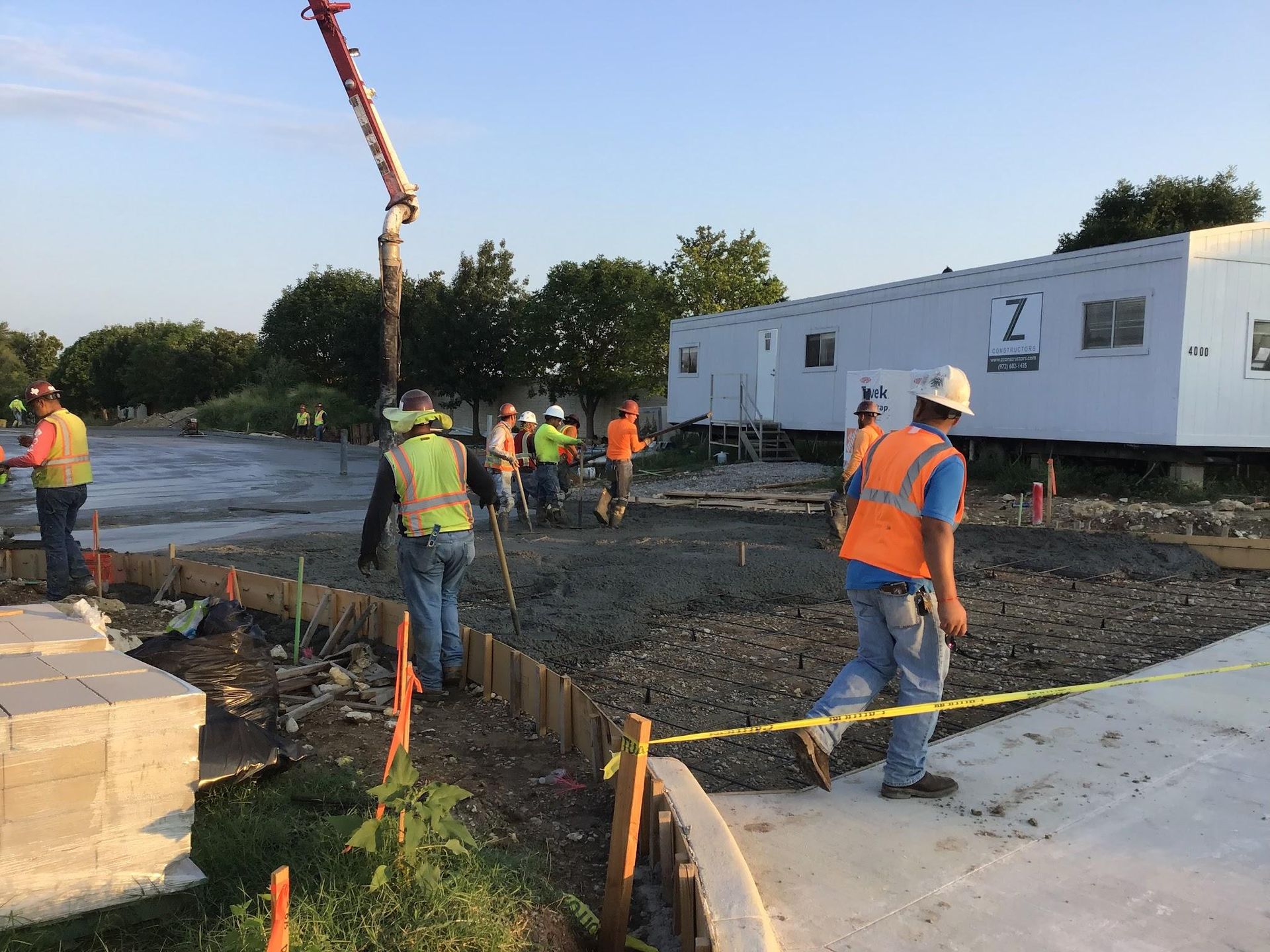 Construction workers pouring concrete at a work site. Several wear safety vests and hard hats near a trailer.