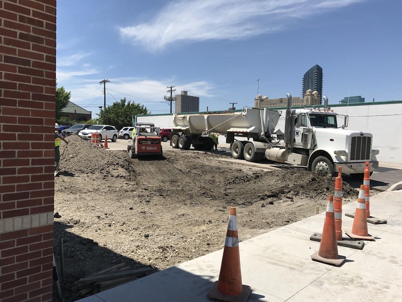 Construction site with dump truck and skid steer; dirt pile on pavement, orange cones.