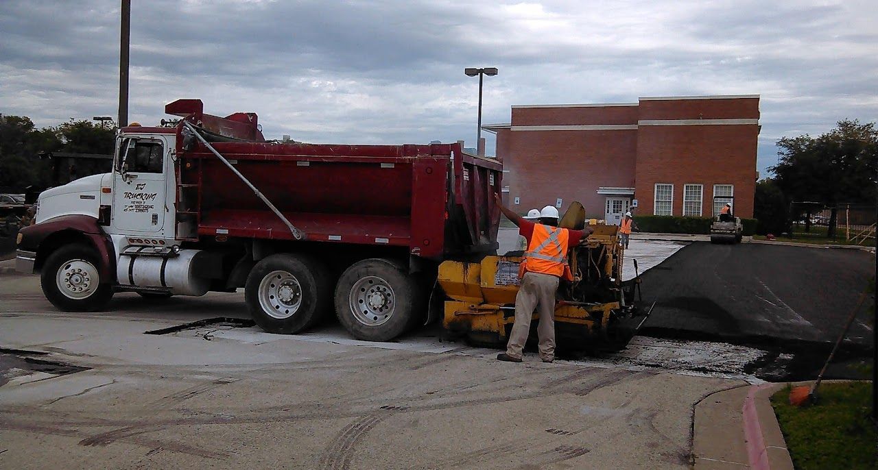 Dump truck unloading asphalt into paving machine at construction site. Worker in orange vest.