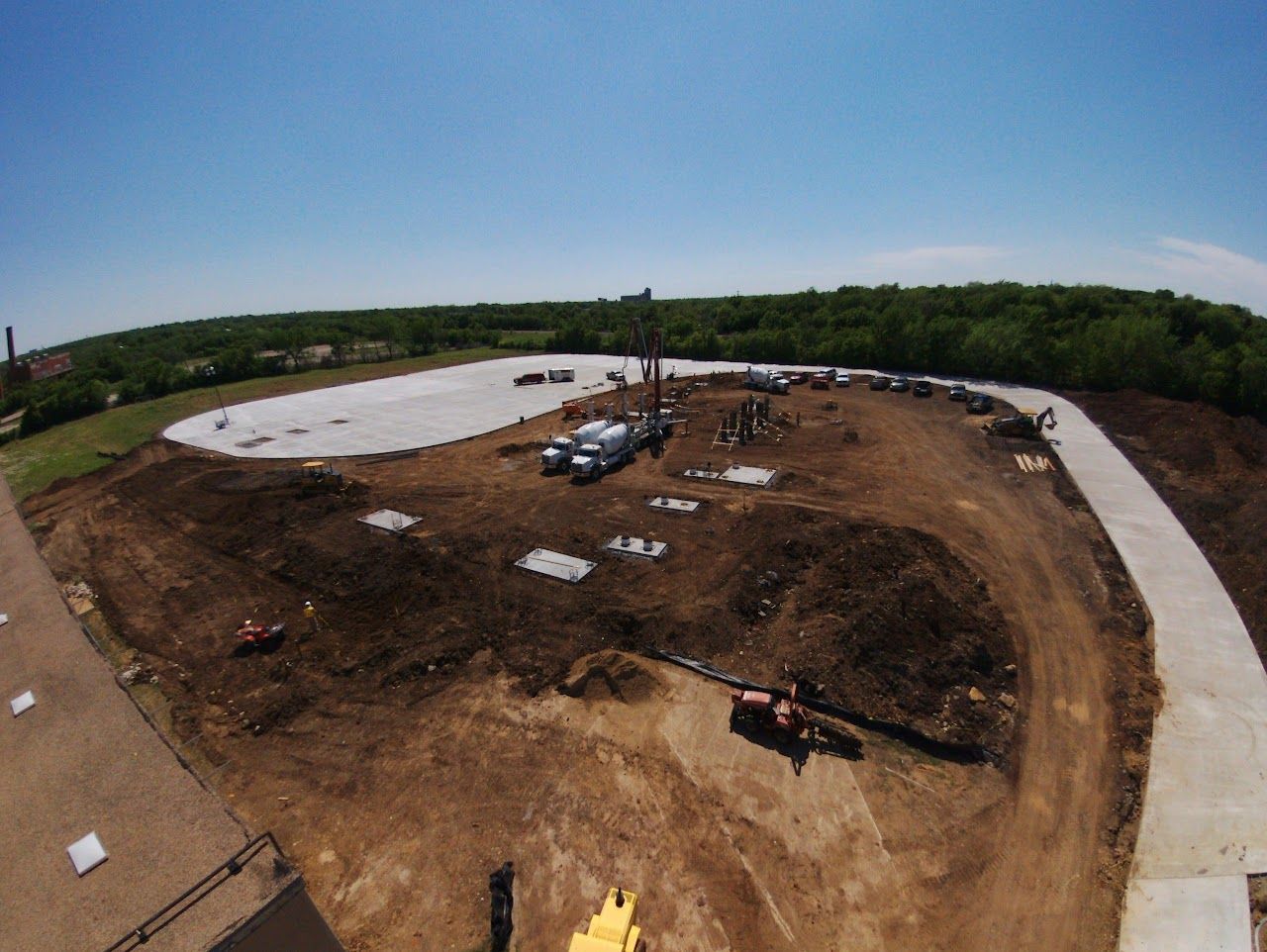 Construction site: earth-moving equipment surrounds a concrete structure. A paved path borders the site, trees in the background.