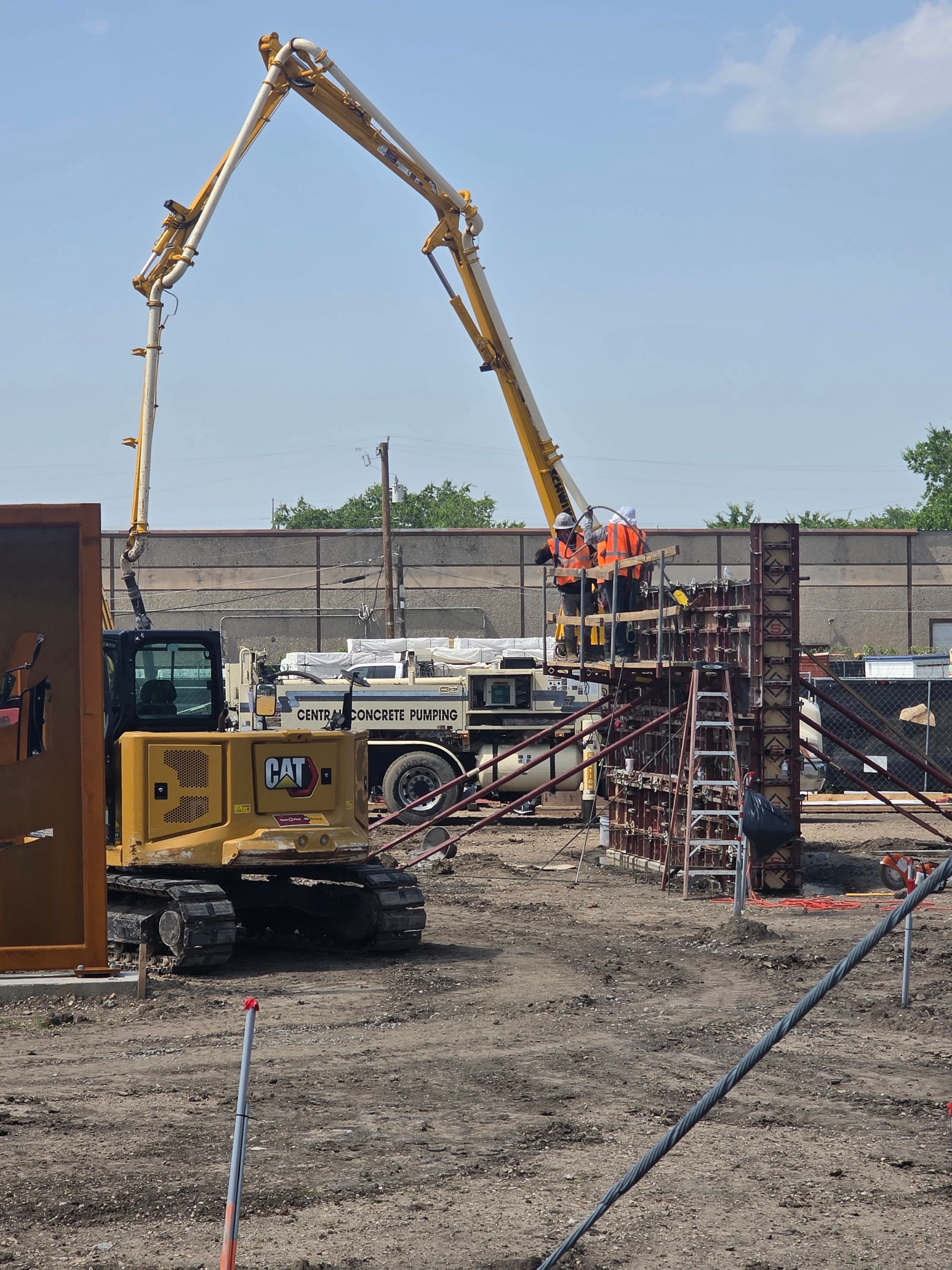 Construction site with a yellow concrete pump truck extending a boom over a wall, workers on scaffolding.