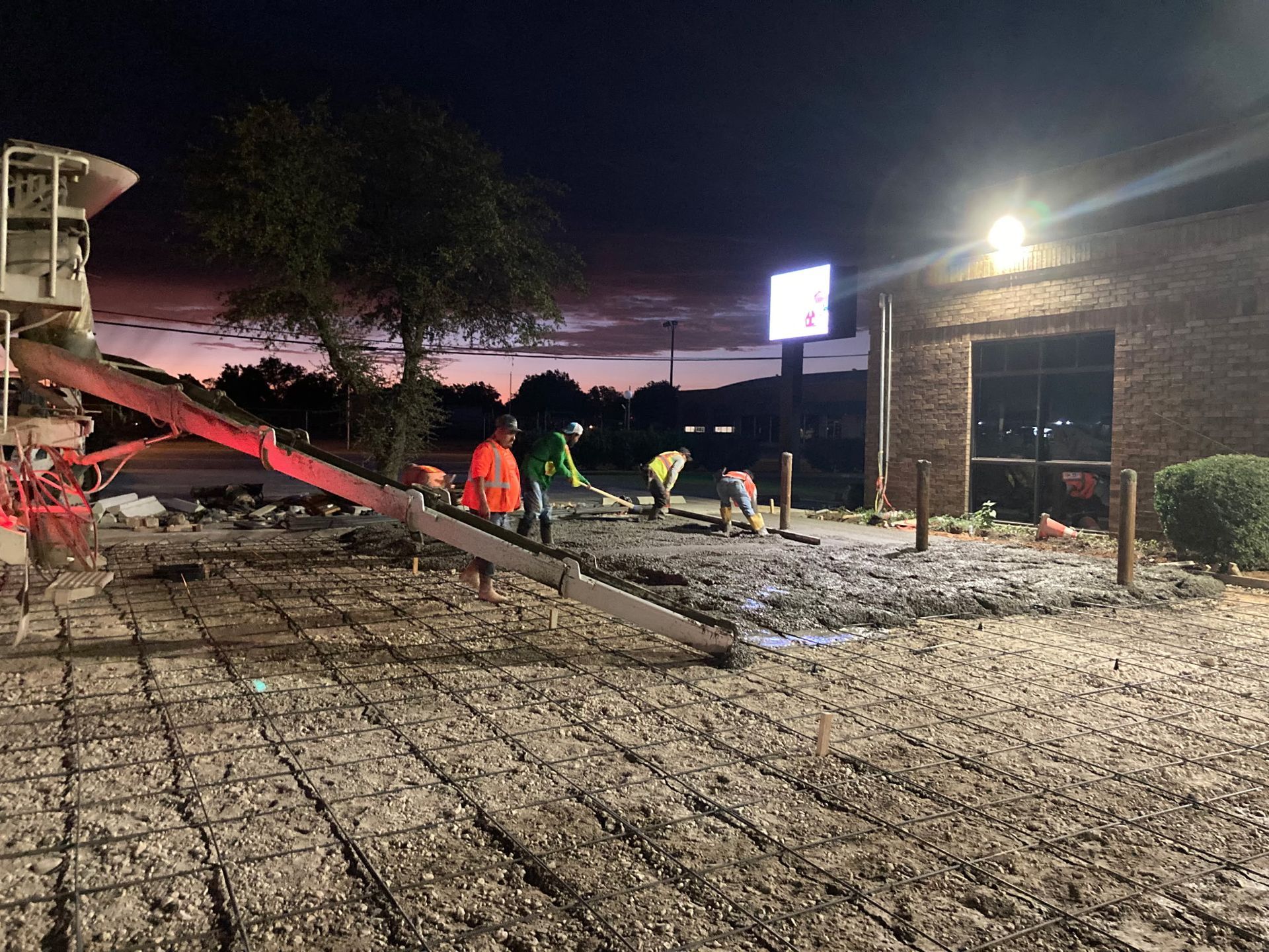 Workers paving a gravel courtyard at dusk beside a brick building, with red equipment and bright lights.