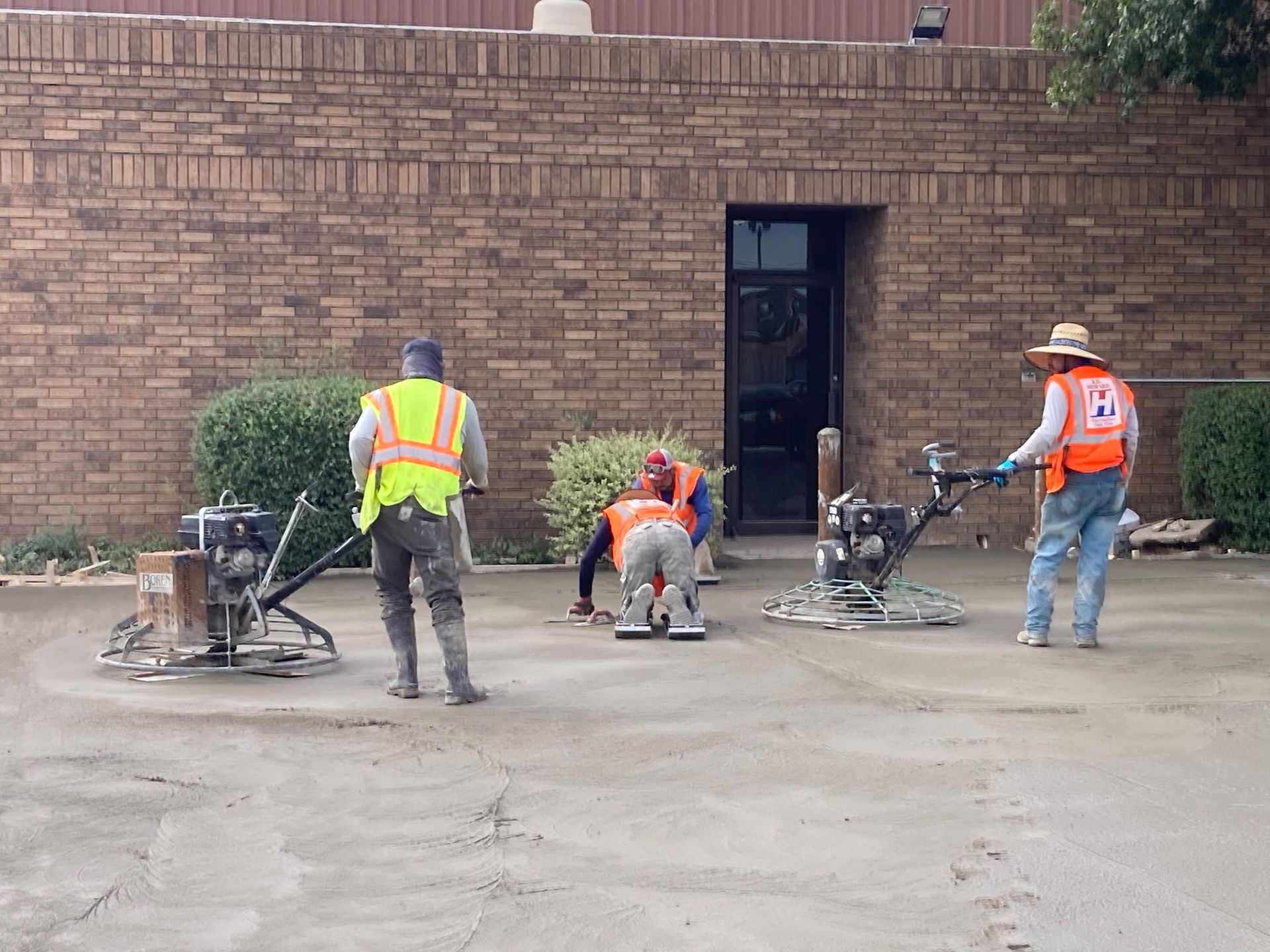 Road workers in orange vests repairing pavement beside a brick wall.