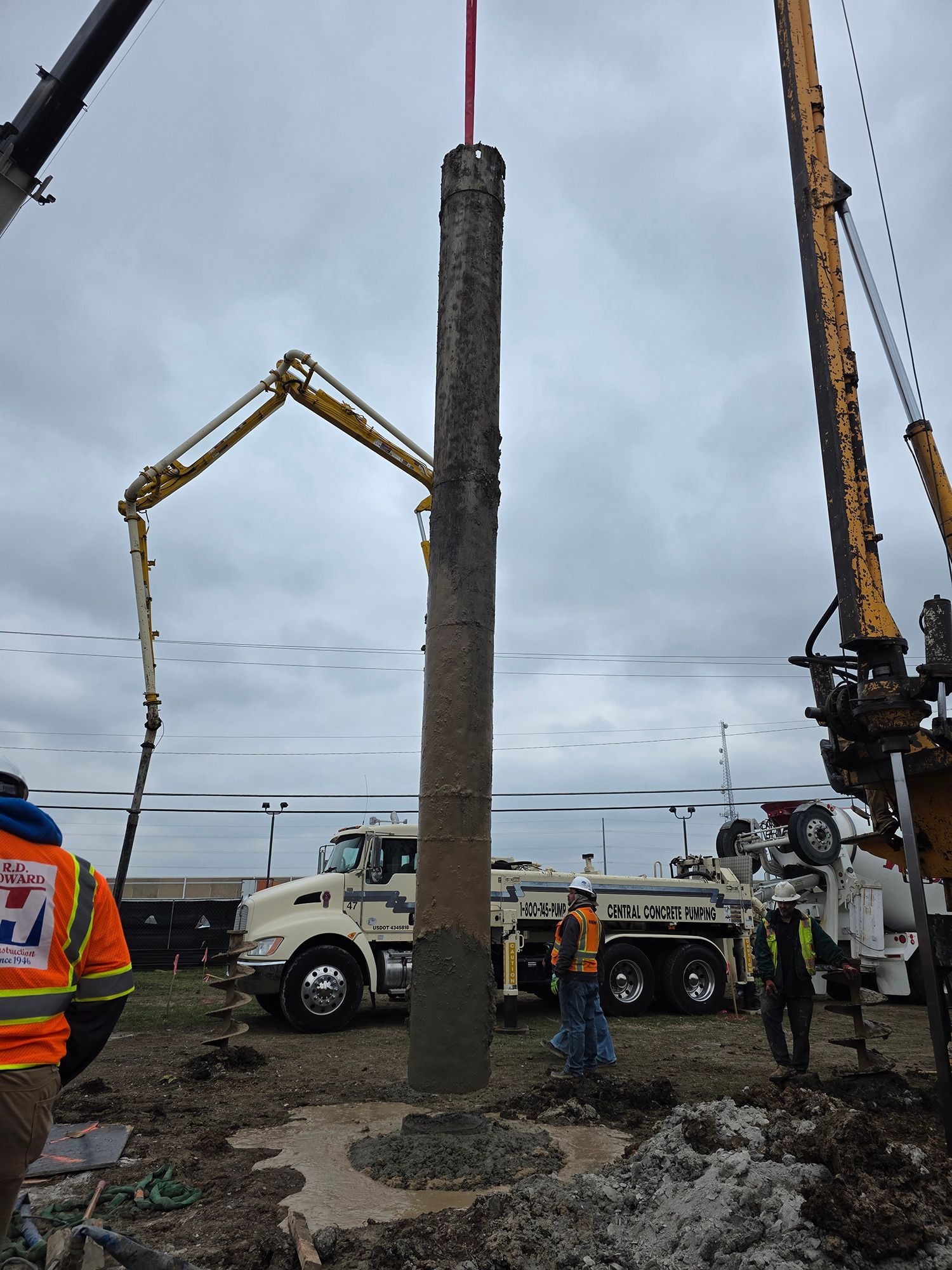 Construction crew lifting a tall concrete pile beside cranes on a muddy worksite near water