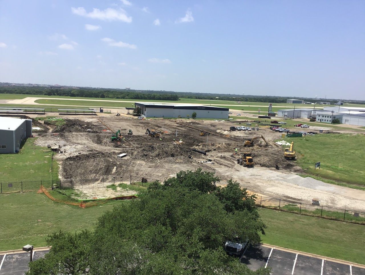 Construction site with heavy machinery, dirt piles, and a partially constructed building on a grassy area, under a bright sky.