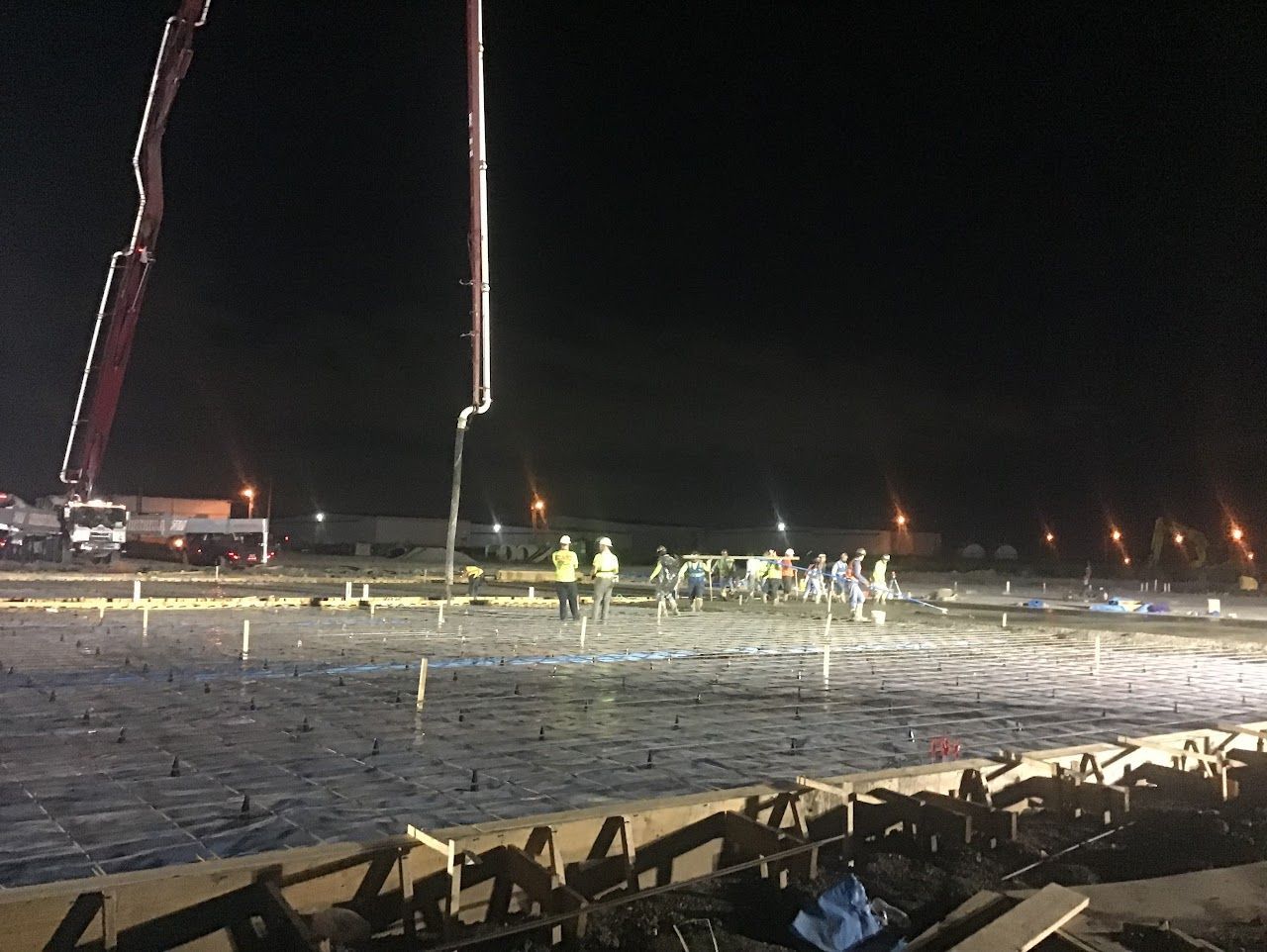 Construction workers pouring concrete at night, using a boom pump and working on a large flat surface.