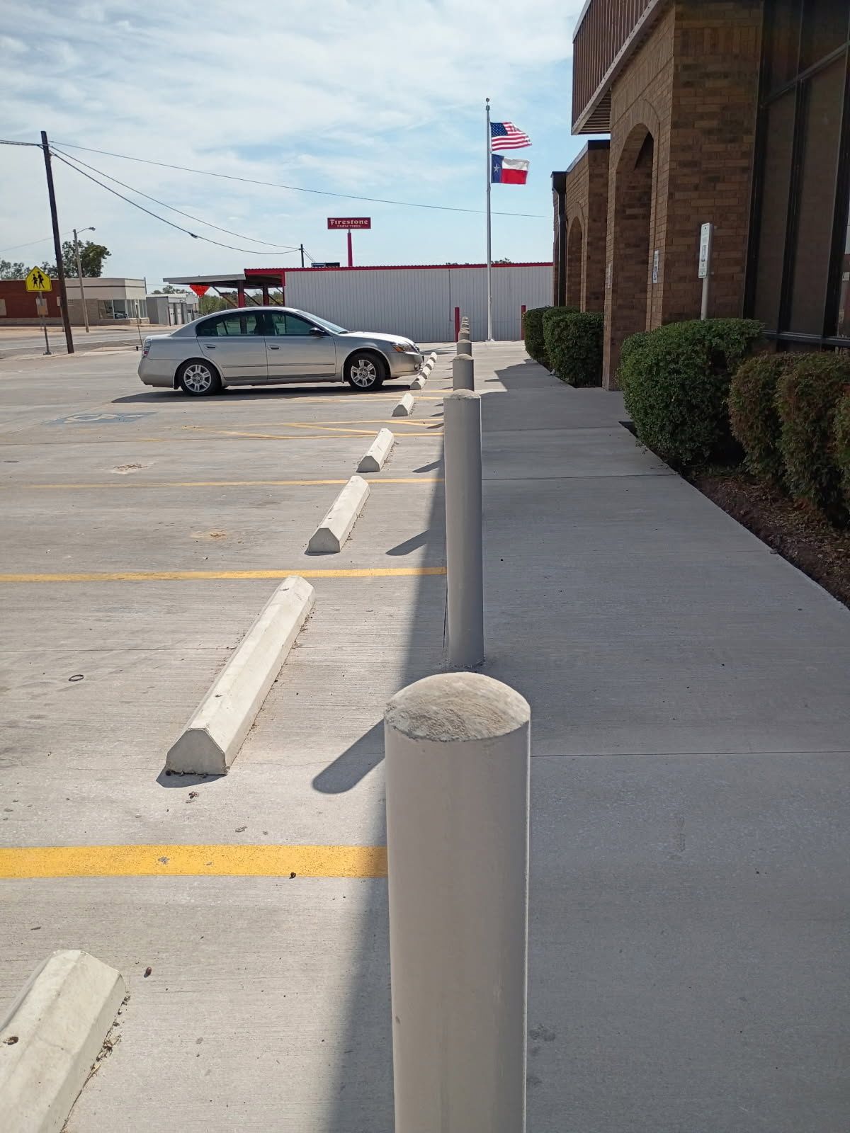 Parking lot beside a brick building, with concrete bollards, a parked car, and an American flag in the distance.