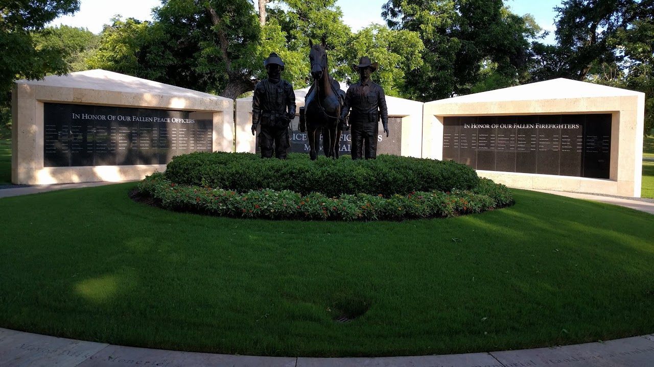Bronze statue of three figures on horseback, flanked by two memorial walls in a park setting.