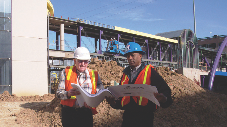 Two construction workers in vests and hard hats review blueprints at a construction site.