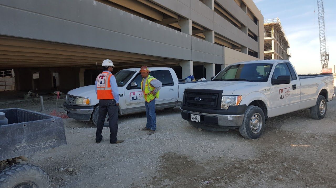 Two men in work vests converse near two white pickup trucks at a construction site.