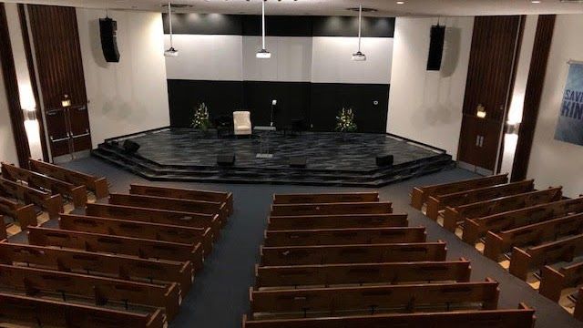 Interior view of a church sanctuary with rows of pews facing a stage. Dark stage, wood paneling, speakers, and a microphone.