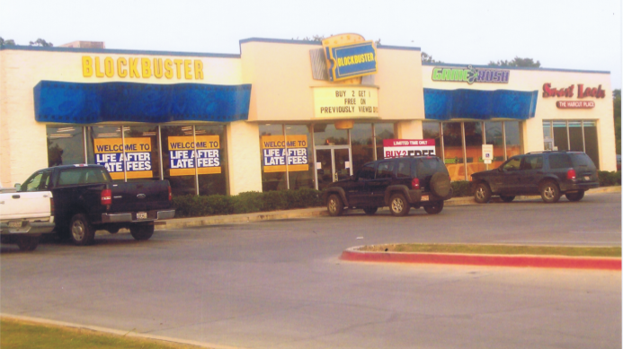Blockbuster video store with sale signs in the windows. Black vehicles parked in front.