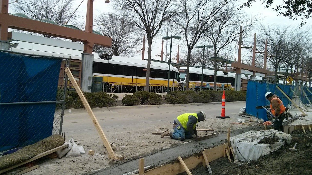 Construction workers pouring concrete at a work site with a building, fence, and trees in the background.
