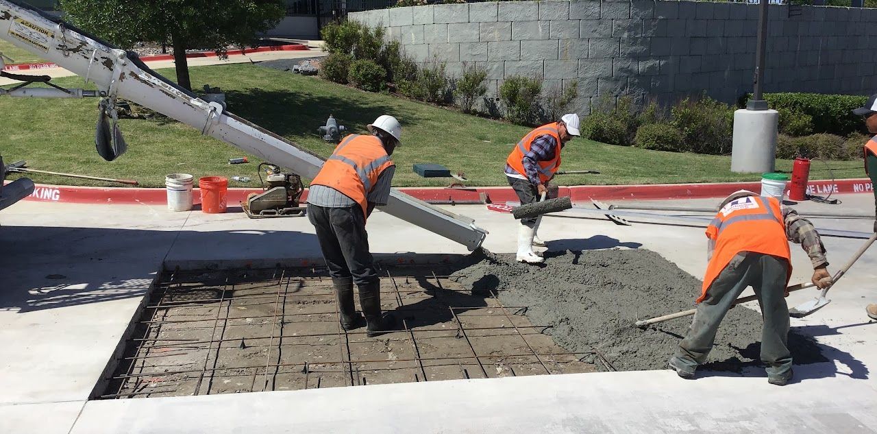 Construction workers in orange vests and hard hats pouring concrete on a sidewalk with a pump truck.