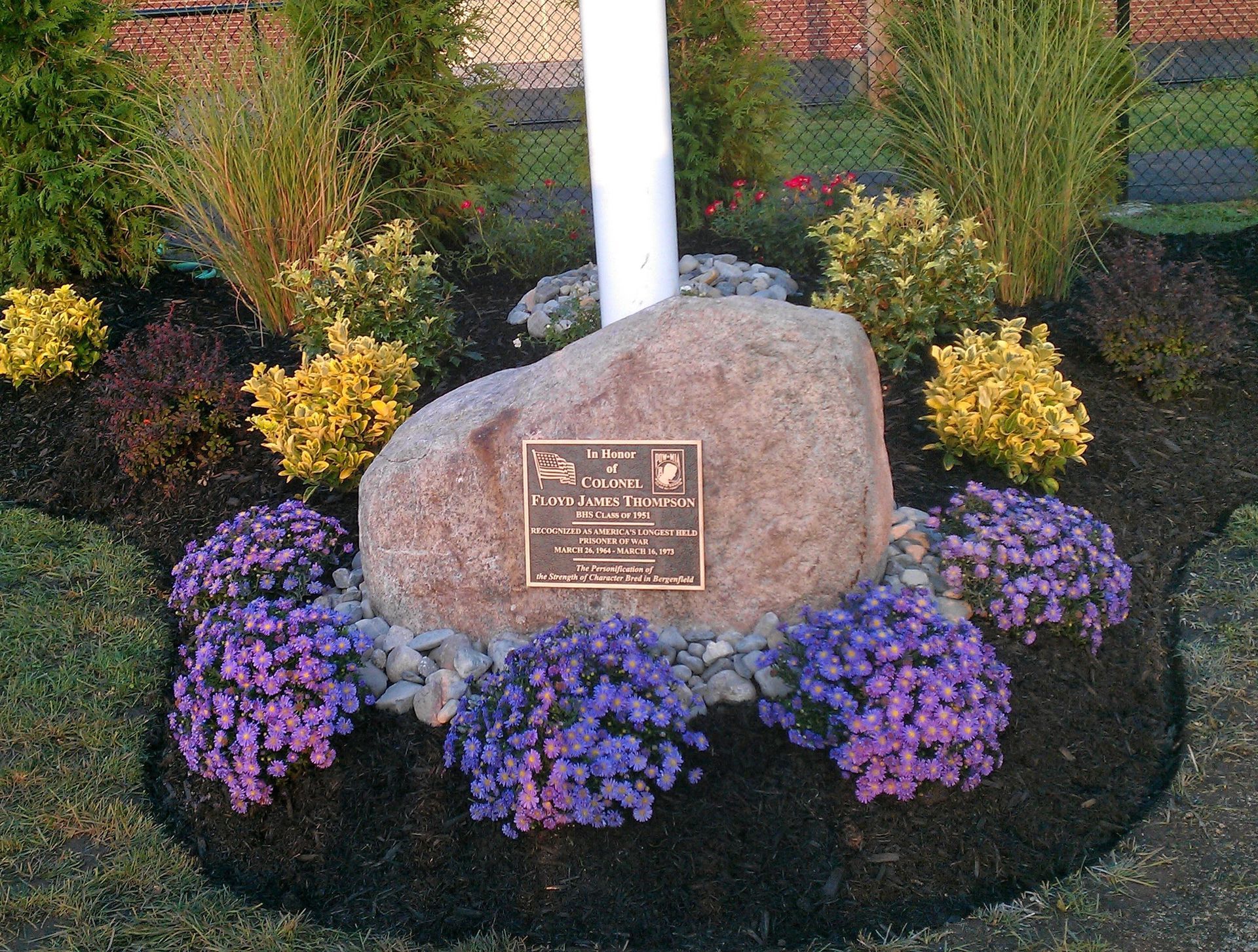 Monument with plaque, surrounded by flowers and greenery, at the base of a flagpole