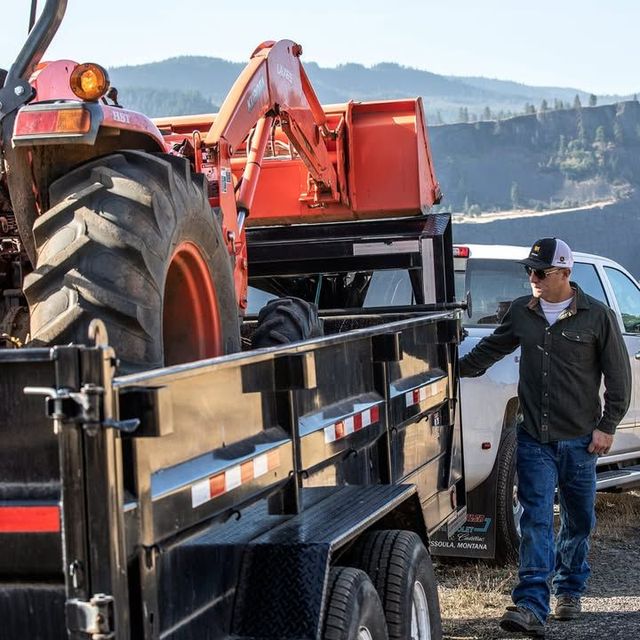 Man near trailer with tractor and equipment, outdoors with hills.