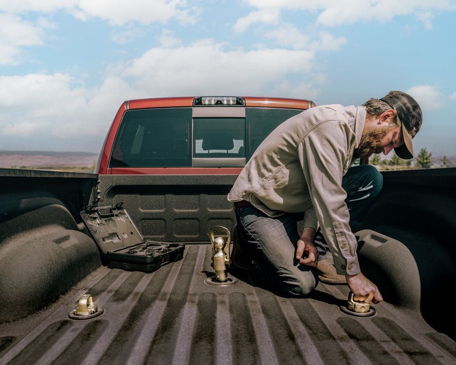 Man installing hitch components in a red pickup truck bed. Outdoors, toolbox nearby, sunny.