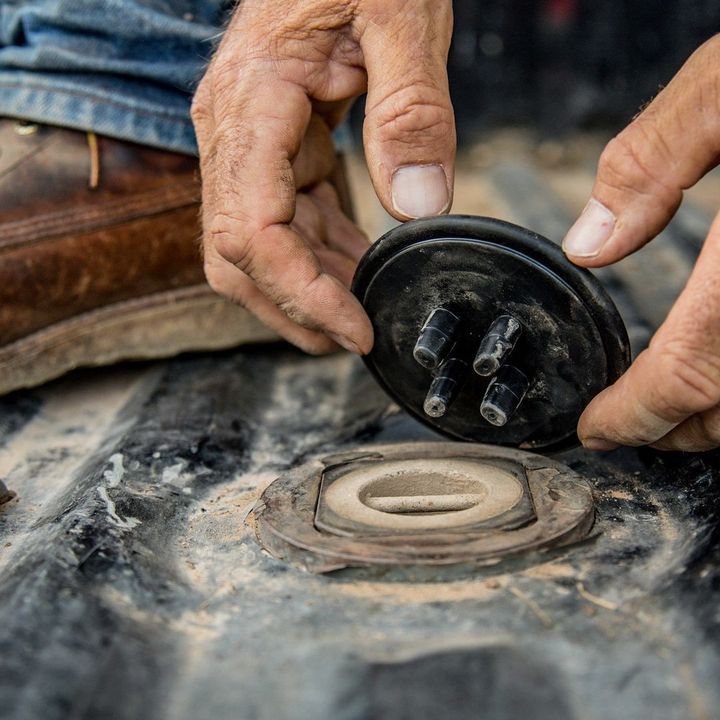 Hands holding a black cap over a fuel tank opening; brown work boot visible.