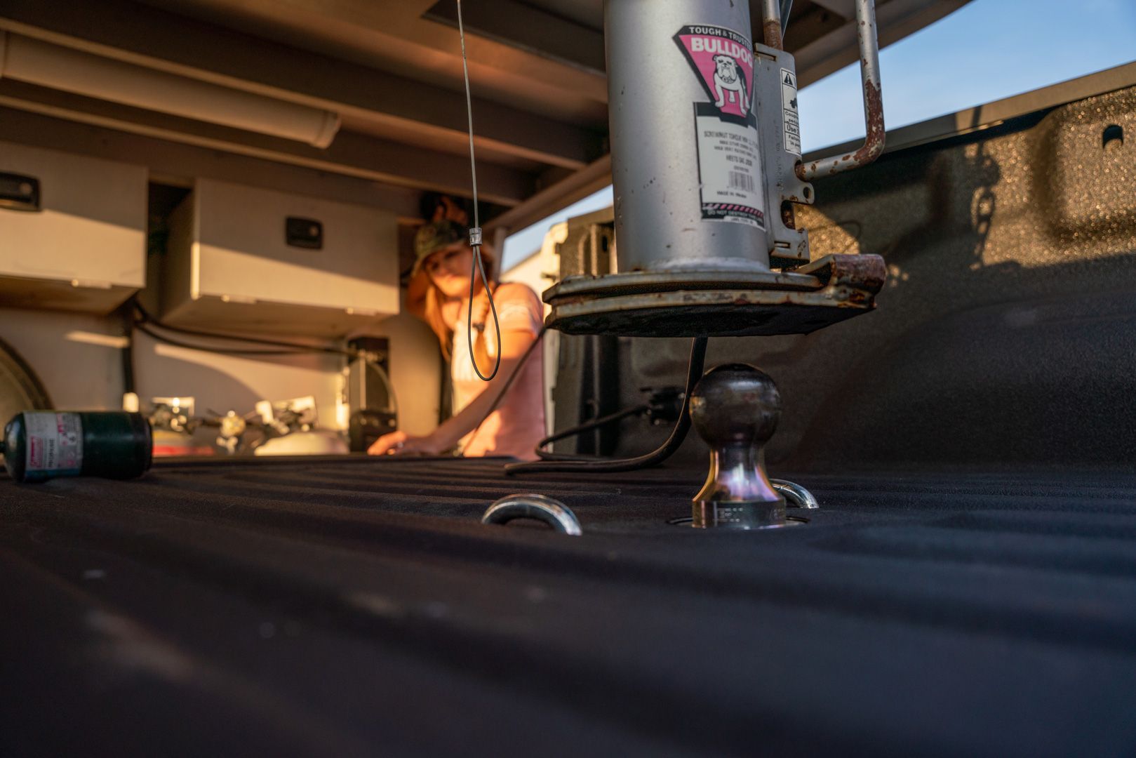 Woman reaching under a truck bed, near a hitch and propane tank, lit by sunlight.