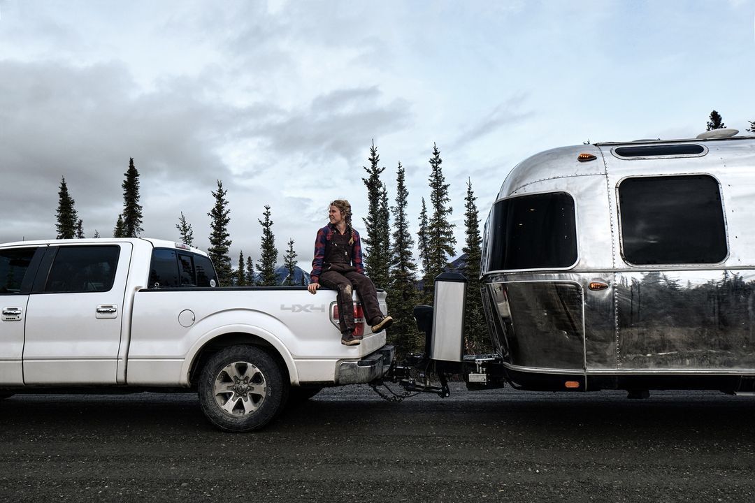 Woman sitting on the tailgate of a white pickup truck, towing a silver Airstream trailer in a mountainous setting.