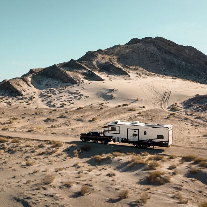 Truck towing a large white and black RV on a dirt road in a desert landscape with a mountain backdrop.
