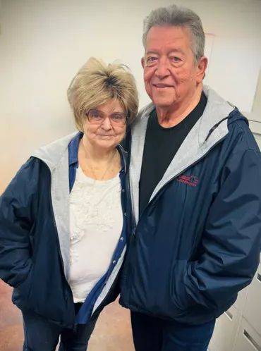 Couple stands closely together, wearing matching blue jackets with hooded linings. Man smiles, woman has neutral expression.