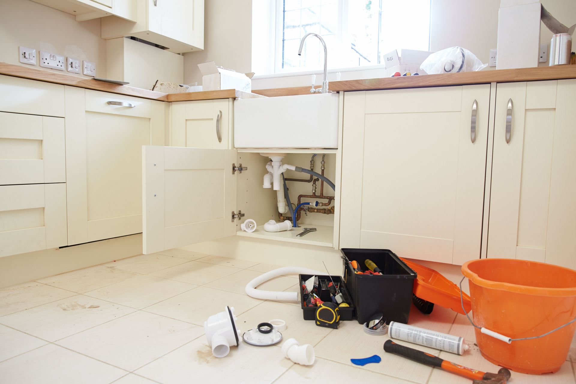 Kitchen with open cabinet, exposed plumbing, tools, and orange bucket on the floor.