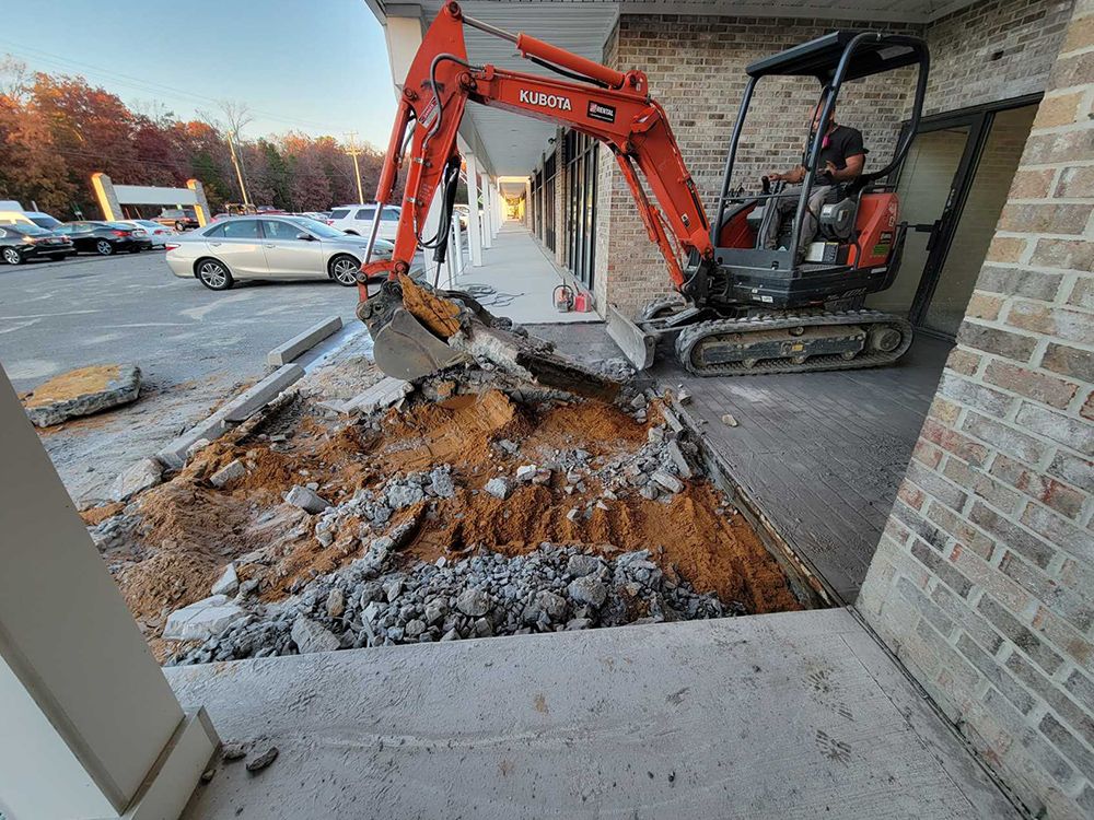 A small excavator is digging a hole in the ground in front of a brick building.