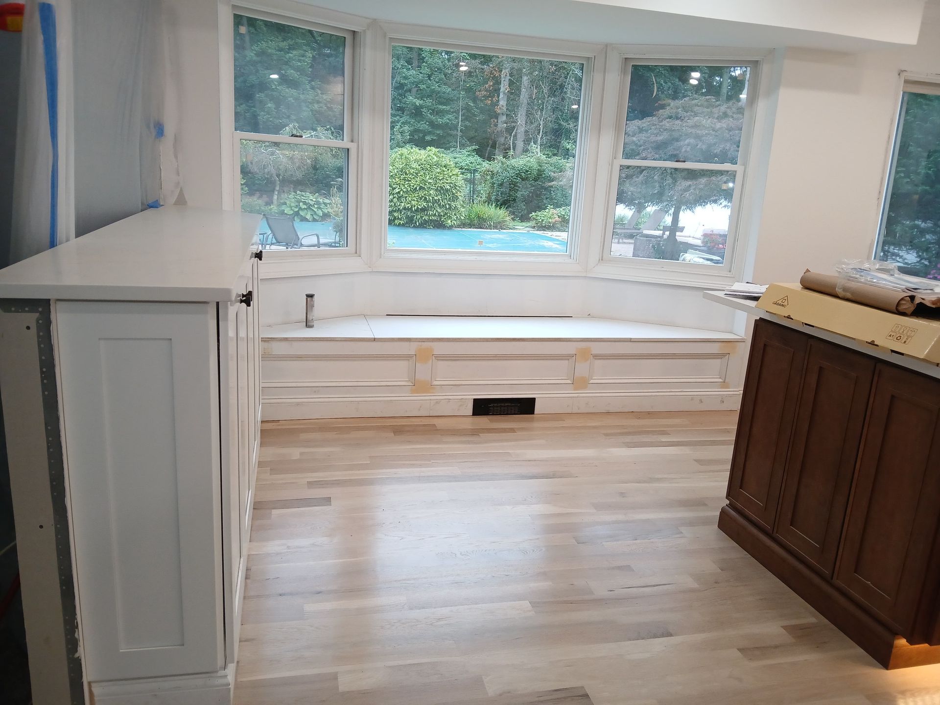 A room with a bay window, cabinets, and light wood floors. The windows overlook greenery.