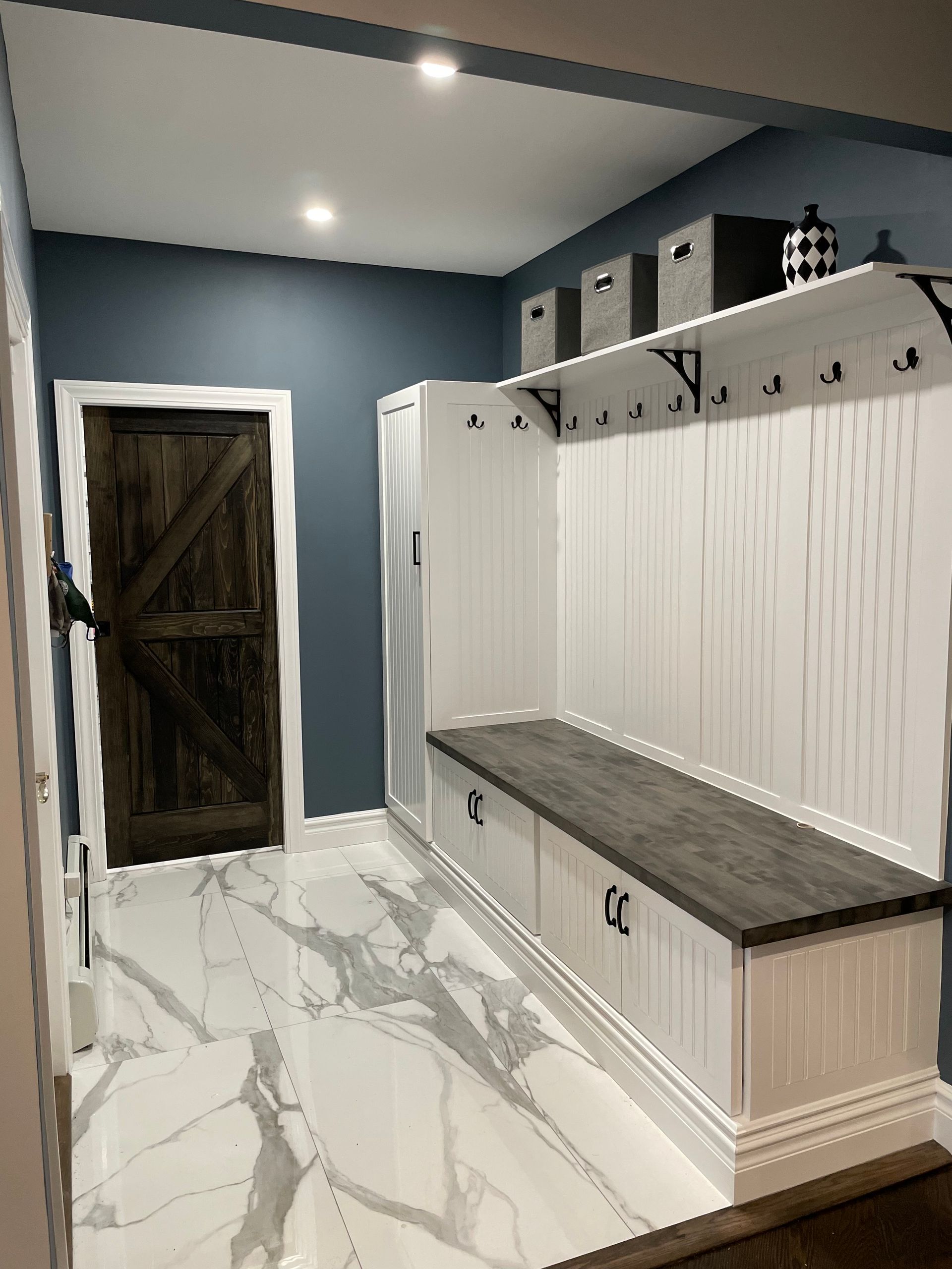 Mudroom with white cabinets, wood bench, hooks, and gray-veined flooring. 
