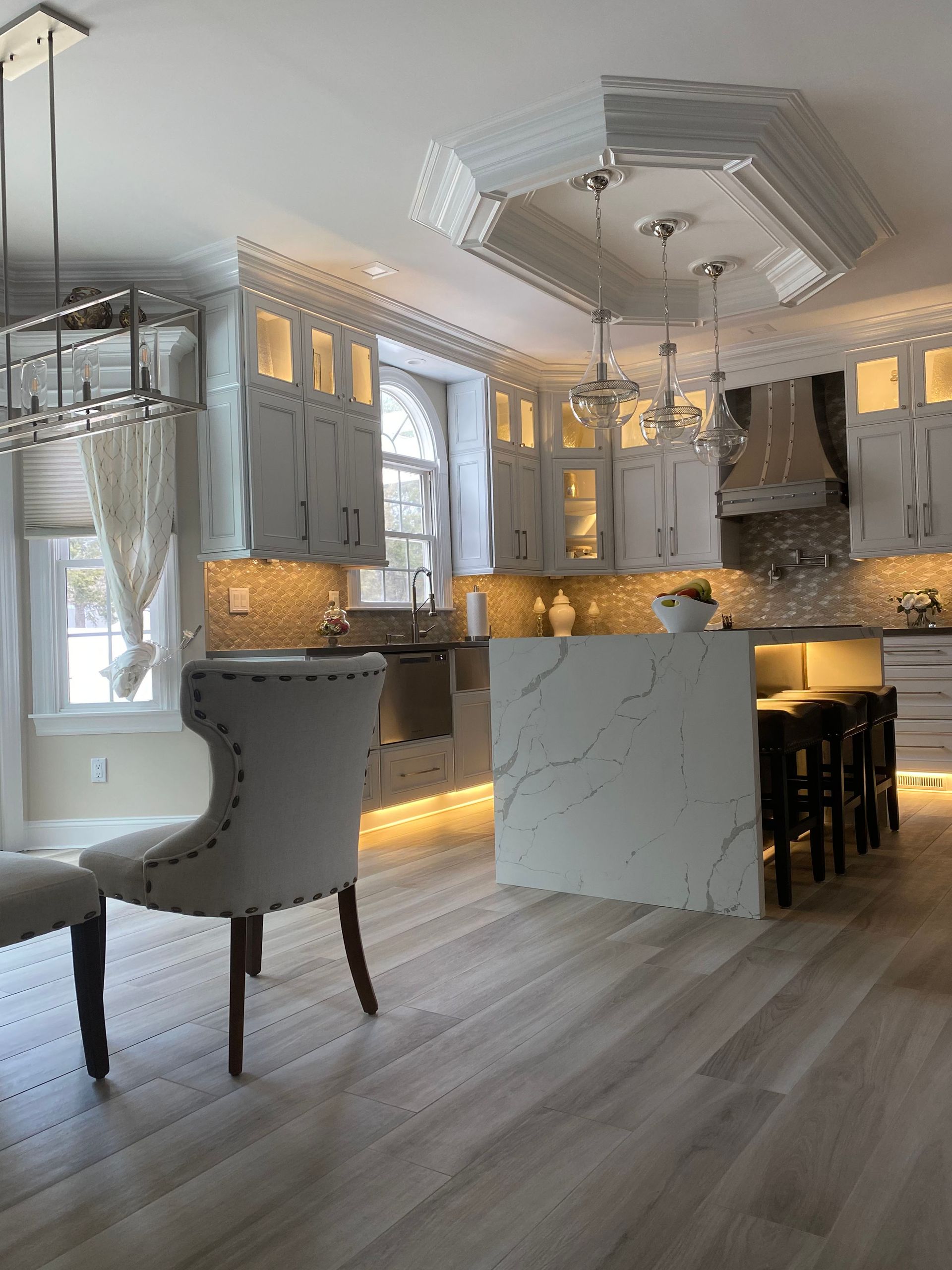 Elegant white kitchen with island, cabinets, and a dining area, featuring modern lighting and light wood floors.